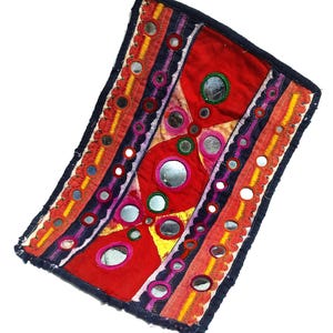 May include: A rectangular textile piece featuring a vibrant red and yellow fabric base with intricate embroidery in purple and orange. The surface is decorated with numerous small circular mirrors framed by colorful stitching and dark blue borders.