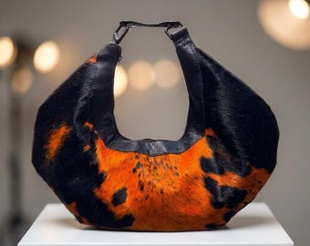Op de afbeelding: Een halvemaanvormige hobo-tas gemaakt van koeienhuid met een opvallend patroon van zwart en feloranje bont. De tas is voorzien van een gladde zwarte lederen rand langs de opening aan de bovenzijde en een centrale lederen handgreepverbinding.