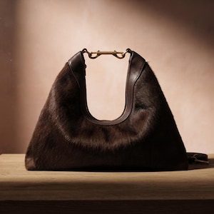 Può includere: Una borsa a mano stile hobo in materiale peloso testurizzato color marrone scuro. La borsa presenta una caratteristica apertura superiore a forma di U, fissata da una chiusura in metallo color ottone che unisce i manici.