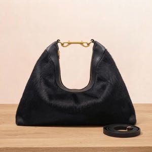 May include: A black hobo bag with a textured hair-on-hide finish, featuring a smooth black leather trim along the top opening and a gold-toned metal hardware connector at the center of the handle. A matching black leather shoulder strap lies nearby.