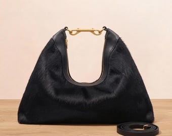May include: A black hobo bag with a textured hair-on-hide finish, featuring a smooth black leather trim along the top opening and a gold-toned metal hardware connector at the center of the handle. A matching black leather shoulder strap lies nearby.