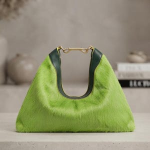 May include: A bright lime green faux fur handbag with a triangular silhouette. The bag features dark forest green leather trim along the top opening and a gold-toned metal hardware connector that secures the two sides of the handle together.