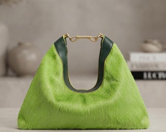 May include: A bright lime green faux fur handbag with a triangular silhouette. The bag features dark forest green leather trim along the top opening and a gold-toned metal hardware connector that secures the two sides of the handle together.
