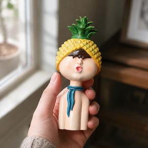 May include: A small figurine of a person with a pale complexion, wearing a yellow pineapple-shaped hat with green leaves on top. The figure has dark hair, a blue scarf around the neck, and a facial expression with pursed lips, rendered in a smooth finish.