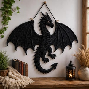 May include: A black crocheted dragon wall hanging featuring large, detailed wings, a long curved tail, and a textured body. The dragon is suspended by a black cord from a small wooden peg, showcasing intricate stitching and a pointed tail tip.