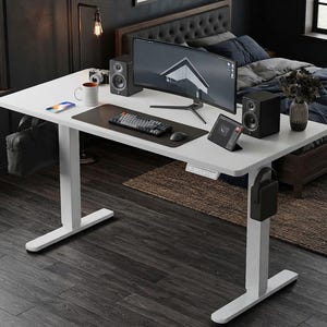 May include: A white standing desk with a rectangular top, featuring a black desk mat, a mechanical keyboard, a computer monitor, two black speakers, a white mug, a smartphone, and a digital control panel mounted on the front edge of the desk surface.