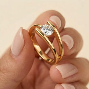 May include: A gold ring featuring a split shank design with a single round brilliant-cut clear gemstone set in the center. The band is polished yellow gold, and the stone is held in place by a tension-style setting that creates an open space.