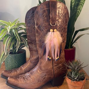 May include: A pair of brown leather cowboy boots featuring intricate white stitching and a pointed toe. A decorative charm made of light pink and brown feathers, wooden beads, and small dark blue beads hangs from the side of the boot shaft.