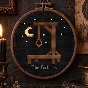 May include: A circular wooden embroidery hoop containing a cross-stitch design on black fabric. The design features a brown wooden gallows structure, a yellow crescent moon, white stars, and the text The Gallows stitched in white thread.