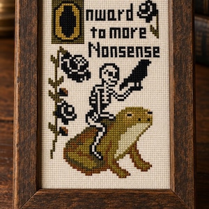 May include: A cross-stitch embroidery in a dark brown wooden frame featuring a skeleton riding a large green frog, holding a black bird. The design includes black flowers and the text "Onward to more Nonsense" stitched in black on a cream fabric.