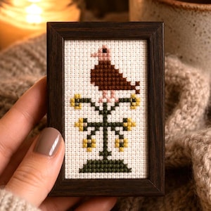 May include: A small rectangular dark wood frame containing a cross-stitch embroidery on white fabric. The design features a brown bird with a light pink head perched atop a dark green metal-style stand decorated with small yellow floral accents.