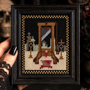 May include: A framed cross-stitch embroidery featuring a brown wooden guillotine with a silver blade, set against a black background with a white snowy ground, two golden crosses, and a decorative gold and black checkered border frame.