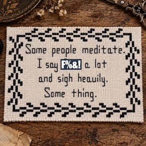 May include: A rectangular cross-stitch sampler on cream-coloured fabric featuring a black stitched border and the text: Some people meditate. I say F%&! a lot and sigh heavily. Some thing. The embroidery is centred with clear black lettering.