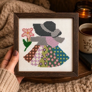 Könnte beinhalten: Eine quadratisch gerahmte Kreuzstichstickerei, die eine Figur mit einem dunkelgrauen Hut und einem mehrfarbigen Patchwork-Kleid zeigt. Die Figur hält eine rosa Blume mit grünen Blättern. Das Motiv ist auf weißem Stoff gestickt.