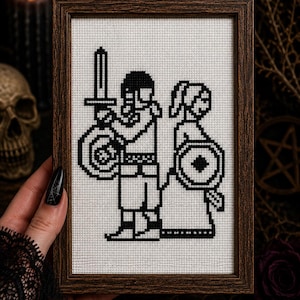 May include: A framed cross-stitch embroidery featuring a black pixelated design on white fabric. The artwork depicts two figures standing side by side, each holding a circular shield. The figure on the left holds a sword pointing upward.