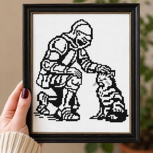 May include: A black and white cross-stitch embroidery featuring a pixelated design of a knight in armor kneeling and petting a cat. The artwork is displayed within a simple black rectangular frame with a thin gold inner border detail.