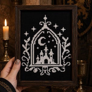 Può includere: Una cornice rettangolare in legno scuro contenente un ricamo a punto croce su tessuto nero. Il disegno in filo bianco raffigura la sagoma di un castello all'interno di un arco a sesto acuto, sormontato da una mezzaluna, stelle e motivi floreali.