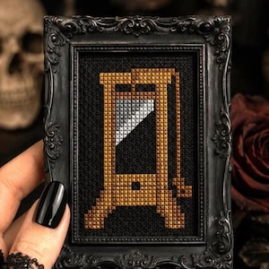 May include: A cross-stitch embroidery featuring a golden-brown guillotine with a silver blade, set against a solid black fabric background. The piece is encased in a small, ornate, matte black rectangular frame with intricate scrollwork detailing.