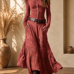 Pode incluir: Uma blusa de manga comprida com botões num tom vermelho terracota suave, combinada com uma saia comprida a condizer com painéis axadrezados integrados e bolsos laterais. Um cinto de pele castanho escuro com fivela circular ajusta a cintura.