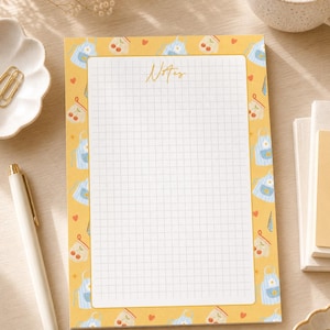 May include: A rectangular notepad with a yellow border featuring a repeating pattern of blue striped aprons, cherry jars, and small red hearts. The centre contains a white grid section with the word Notes printed in a cursive script at the top.
