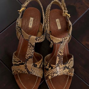 May include: A pair of T-strap sandals featuring a snakeskin pattern in shades of tan, brown, and black. The sandals have brown leather insoles with a gold-tone buckle closure on the ankle strap and a visible Miu Miu brand label on the heel area.