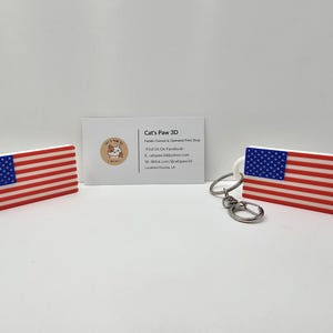 Op de afbeelding: Twee rechthoekige objecten met het ontwerp van de vlag van de Verenigde Staten in rood, wit en blauw. Het ene object is een los stuk, terwijl het andere is bevestigd aan een zilveren metalen sleutelhanger. Een visitekaartje van Cat's Paw 3D staat ertussen.