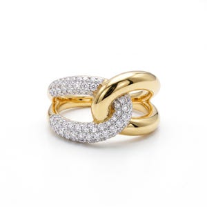 Op de afbeelding: Een gouden ring met een in elkaar grijpend schakelontwerp. De ene kant van de ring is glad gepolijst goud, terwijl de andere kant is ingelegd met talloze kleine, heldere, sprankelende steentjes die het oppervlak van de metalen schakel bedekken.