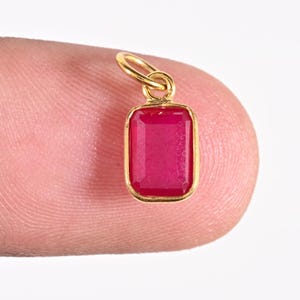 May include: A small rectangular pendant featuring a vibrant pinkish-red gemstone set in a polished gold-toned metal bezel. The pendant includes a small circular gold-toned jump ring at the top for attachment, showcasing a clean and simple design.
