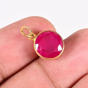 Op de afbeelding: Een ronde hanger met een gefacetteerde, levendige magenta-roze edelsteen in een gladde, goudkleurige metalen zetting. De hanger is voorzien van een klein, rond, goudkleurig verbindingsringetje aan de bovenkant om aan een ketting te hangen.