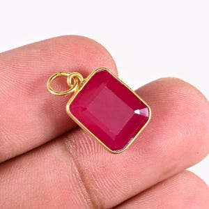 Peut inclure: Un pendentif rectangulaire doté d'une pierre précieuse facettée rose foncé sertie dans une monture en métal doré. Le pendentif comprend un petit anneau circulaire fixé sur le bord supérieur, permettant de le suspendre à une chaîne ou un cordon.
