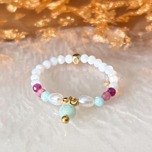 May include: A beaded ring featuring small white round beads, two white freshwater pearls, two faceted magenta beads, two light blue beads, and a central gold-toned metal charm with a dangling light blue bead and a small gold-toned sphere.