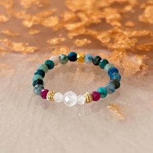 May include: A beaded ring featuring a circular arrangement of small, faceted teal, blue, and dark green beads, accented with two bright magenta beads, a central clear faceted bead, and small gold-toned metallic spacers throughout the band.