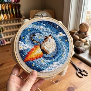 May include: A round wooden embroidery hoop containing a fabric piece with a hand-stitched design of a calico cat flying through a swirling blue night sky. The cat has a fiery orange and yellow rocket trail behind it, surrounded by small white daisies.