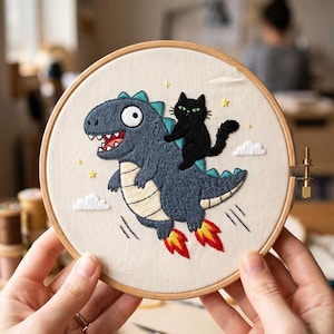 May include: A circular wooden embroidery hoop containing a fabric panel with a hand-stitched design of a grey dinosaur with a cream-colored belly flying through the air with red and yellow flames, carrying a small black cat on its back.