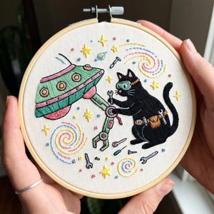 May include: A circular wooden embroidery hoop containing a fabric panel with a hand-stitched design of a black cat wearing goggles and a tool belt, repairing a green and pink UFO with a wrench, surrounded by colorful embroidered stars and galaxies.