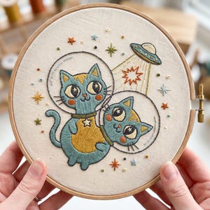 Puede incluir: Un bastidor de bordado con una tela color hueso que presenta un diseño cosido a mano de dos gatos azules con cascos espaciales de cristal. Arriba, un pequeño platillo volante emite un rayo de luz amarilla hacia una estrella. La escena incluye estrellas bordadas de colores y pequeñas cuentas dispersas por la tela.