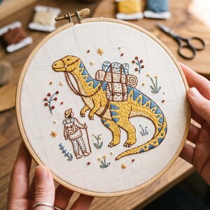 Pode incluir: Um bastidor de bordado circular de madeira contendo tecido branco com um desenho bordado à mão de um dinossauro amarelo e azul usando uma sela com mochilas, acompanhado por uma pequena figura com chapéu e mochila entre motivos florais.