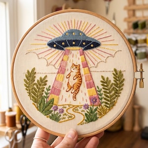 May include: A circular wooden embroidery hoop containing a fabric panel with a hand-stitched design of a blue UFO beaming down a light onto an orange tabby cat. The scene includes green foliage, small purple flowers, and yellow sunburst rays.