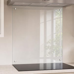 May include: A rectangular, clear glass kitchen backsplash panel mounted vertically behind a black stovetop. The panel is secured to the wall with four circular, metallic silver hardware fasteners positioned at each of the four corners.