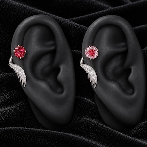 Puede incluir: Dos pendientes tipo ear cuff sobre modelos de oreja negros. Cada pieza presenta un perno de piedra roja redonda y facetada en la parte superior y un ala plateada curva incrustada con pequeñas piedras blancas brillantes en el borde inferior.