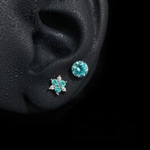 Può includere: Due orecchini a lobo color argento con pietre color verde acqua brillante. Un orecchino è una singola pietra a taglio rotondo, mentre l'altro è un gruppo di tre pietre più piccole a forma di fiore con minuscole pietre trasparenti.
