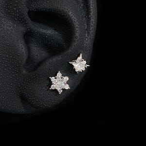 May include: Two silver-toned stud earrings featuring clear, faceted stones. One earring is shaped like a six-pointed snowflake, while the other is a four-pointed star. Both pieces are set with small, sparkling crystals that catch the light.