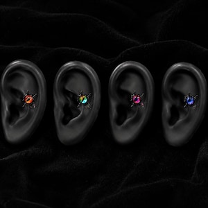 Könnte beinhalten: Vier schwarze Ohrstecker mit einem zentralen Augendesign und vertikaler Pupille. Jeder Stecker zeigt eine andere Irisfarbe: Orange, Grün, Pink und Blau. Die Stecker sind in einen dunklen, stacheligen Metallrahmen gefasst und auf vier schwarzen Ohrmodellen präsentiert.