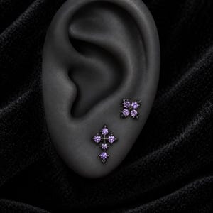May include: Two black metal stud earrings featuring purple round-cut stones. One earring is shaped like a four-pointed star, and the other is shaped like a cross. Both pieces have a dark finish and are set with small, vibrant purple gemstones.