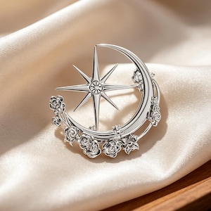 May include: A silver-toned metal brooch featuring a crescent moon shape adorned with small sculpted roses along the outer edge. A prominent eight-pointed star with a central clear crystal accent is positioned within the curve of the moon.