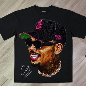 May include: A black short-sleeve t-shirt featuring a large, detailed graphic made of sparkling rhinestones. The design depicts a person wearing a black baseball cap with pink accents, black sunglasses, and a jeweled necklace, with the initials CB.