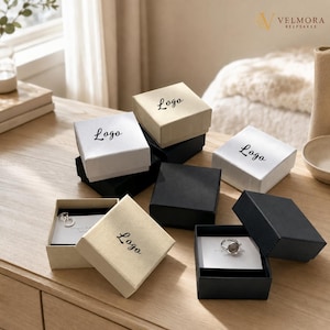 Puede incluir: Una colección de cajas cuadradas para joyería en negro, blanco y beige, cada una con la palabra Logo impresa en la tapa. Algunas cajas están abiertas, revelando anillos de plata sobre tarjetas blancas, mientras que otras permanecen cerradas.
