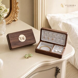 May include: A dark wood jewelry box with a gold butterfly emblem on the lid. The open box features a light gray velvet interior with hooks for necklaces, a compartment for a bracelet, and slots for rings. The text VELMORA KEEPSAKES is visible.