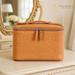 Può includere: Un beauty case rettangolare color cuoio con texture goffrata effetto struzzo, una maniglia superiore e una chiusura a doppia zip argentata. Nell'angolo in alto a destra è presente la scritta VELMORA KEEPSAKES con un piccolo logo.