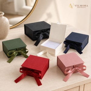 Puede incluir: Una colección de cajas de regalo cuadradas en negro, verde bosque, azul marino, rojo y rosa empolvado, cada una con un cierre de cinta de satén a juego. Una caja color crema está abierta, revelando un inserto interior blanco. El logo Velmora Keepsakes es visible.